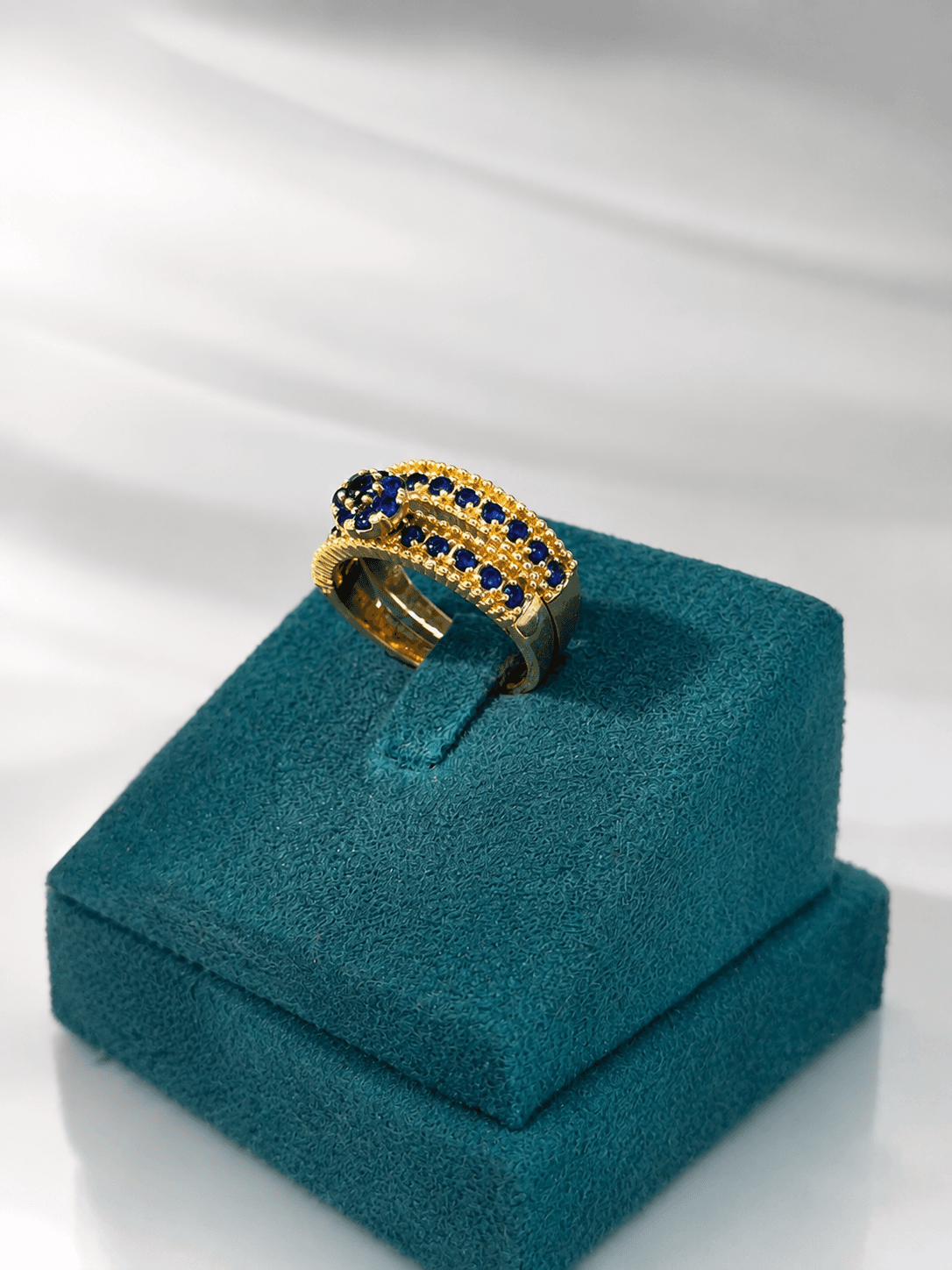 Wide gold band ring with rows of blue gemstones on a teal velvet display stand.