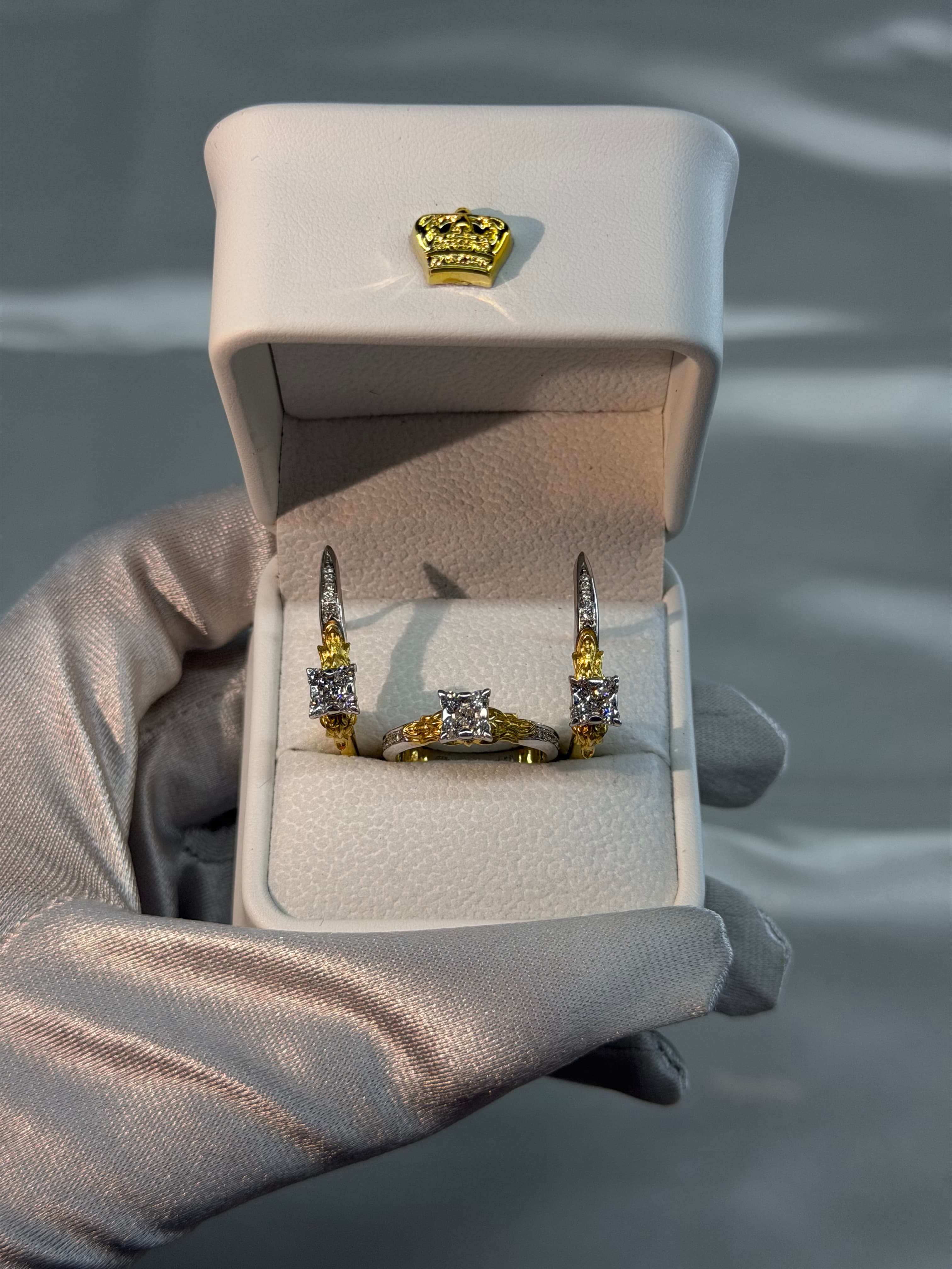 Gloved hand holding an open white box featuring a two-tone gold and diamond jewelry set.